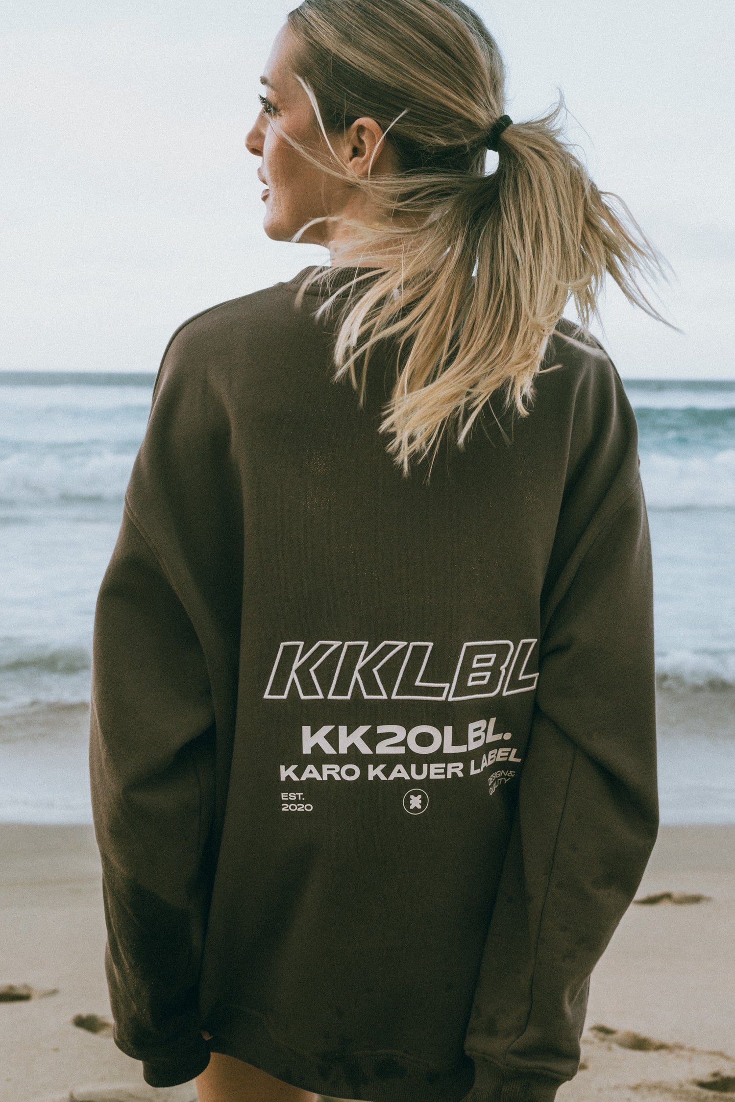 Frau im olivgrünen Oversized Sweater mit weißem KKLBL und KK20LBL Print, Rückansicht