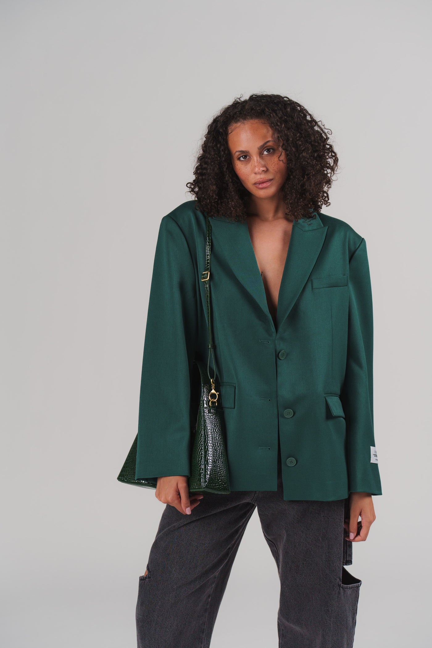 Frau in dunkelgrünem Blazer und grüner Kroko-Handtasche aus der Evergreen Collection mit schwarzen Jeans vor grauem Studio-Hintergrund