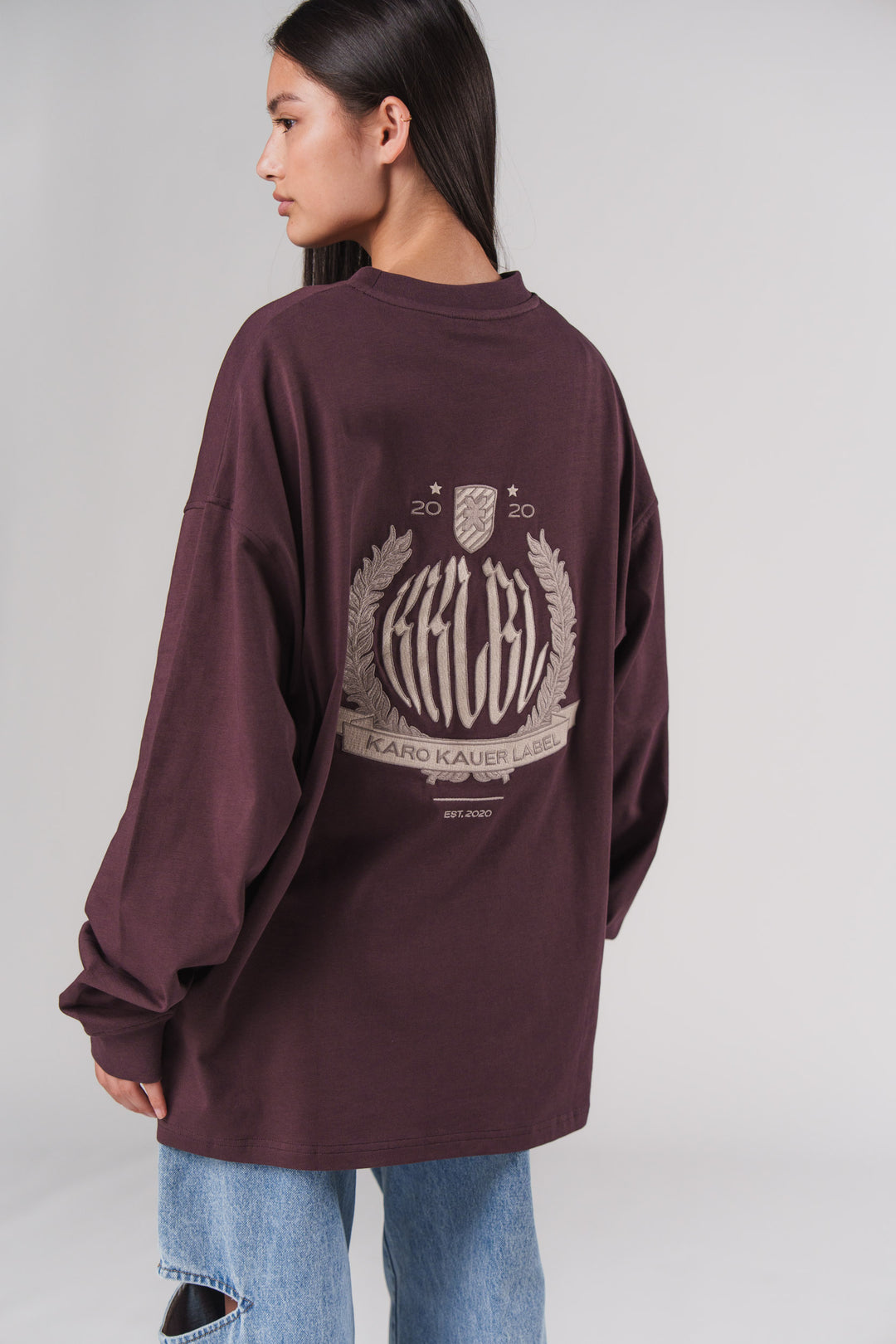 Rückansicht: Frau im pflaumenfarbenen Longsleeve Plum mit KARO KAUER LABEL Wappen-Print