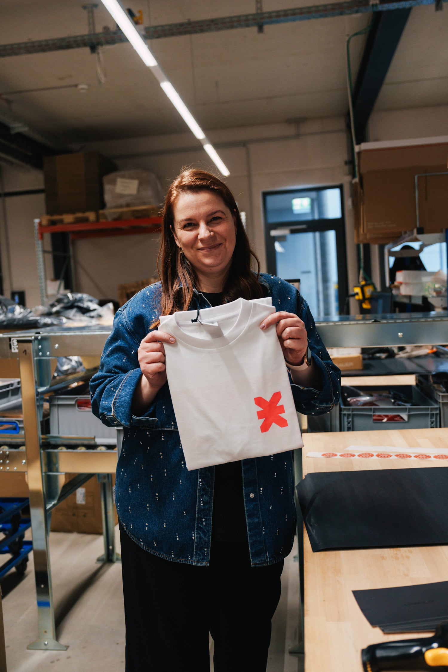 Frau in blauer Jeansjacke hält weißes Kommunity T-Shirt mit neonrotem X-Print
