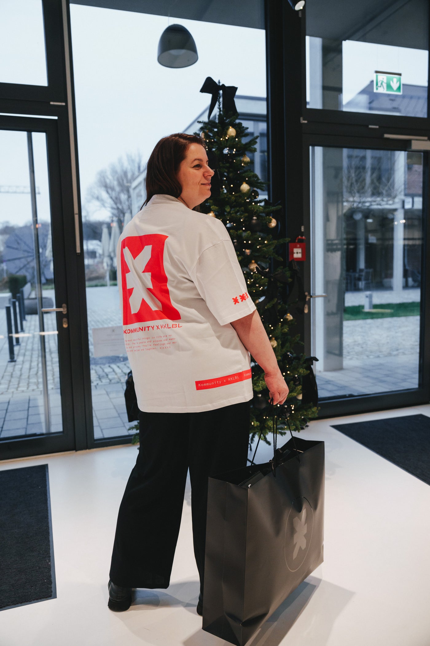 Seitliche Rückansicht: Frau im weißen Kommunity T-Shirt Pink mit neonrotem X-Print und KKLBL Logo