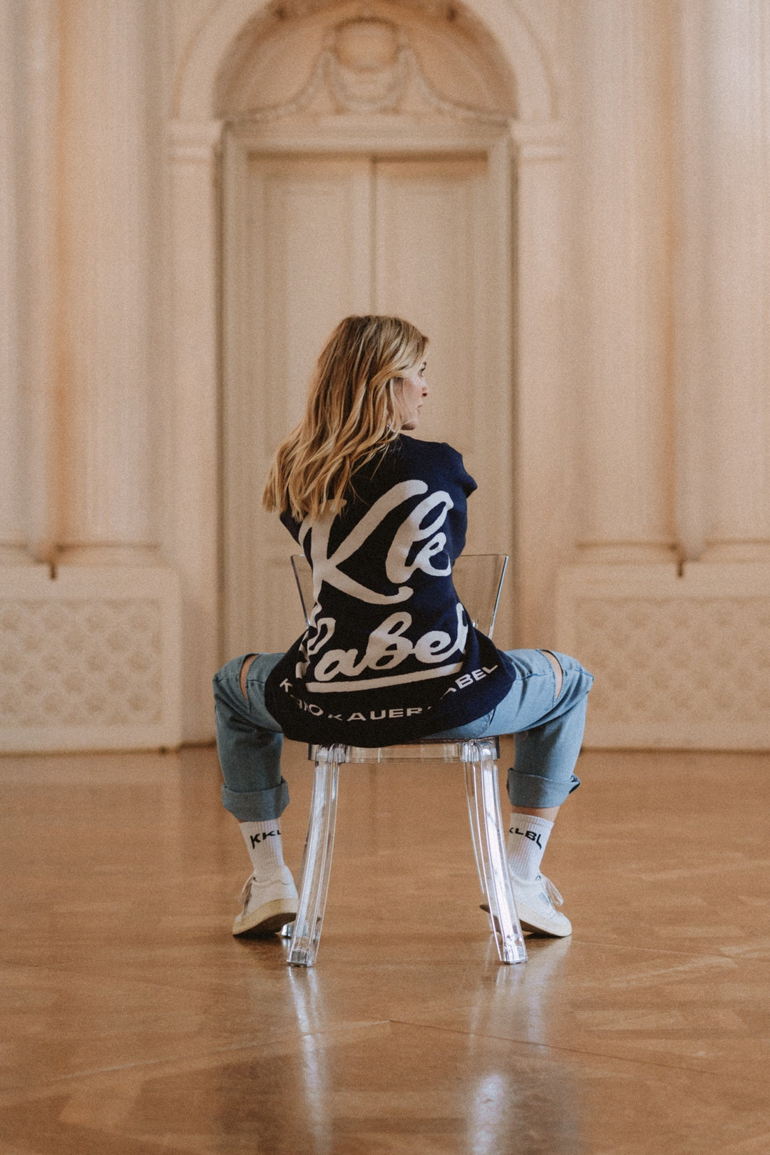 Ganzkörper: Frau im dunkelblauer Knit Sweater KK Label mit weißem KKLBL Print, sitzende Rückansich