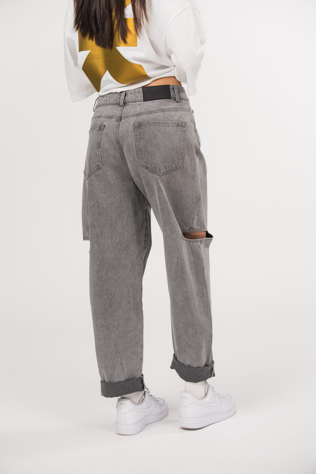 Ein Model, von hinten gezeigt, trägt eine graue Cut-out-Jeans aus der No Hustle Culture Kollektion mit umgeschlagenem Saum, kombiniert mit weißen Sneakern
