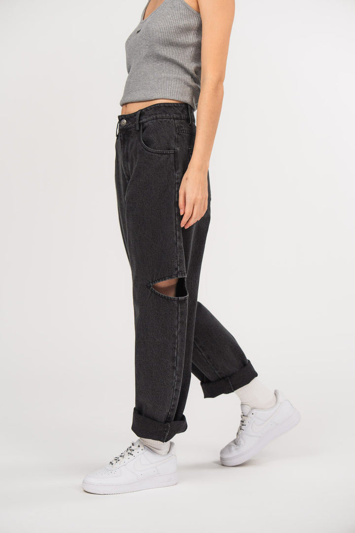 Ein Model trägt eine schwarze Cut-out-Jeans aus der No Hustle Culture Kollektion mit umgeschlagenem Saum, kombiniert mit einem grauen Tanktop, weißen Socken und weißen Sneakern