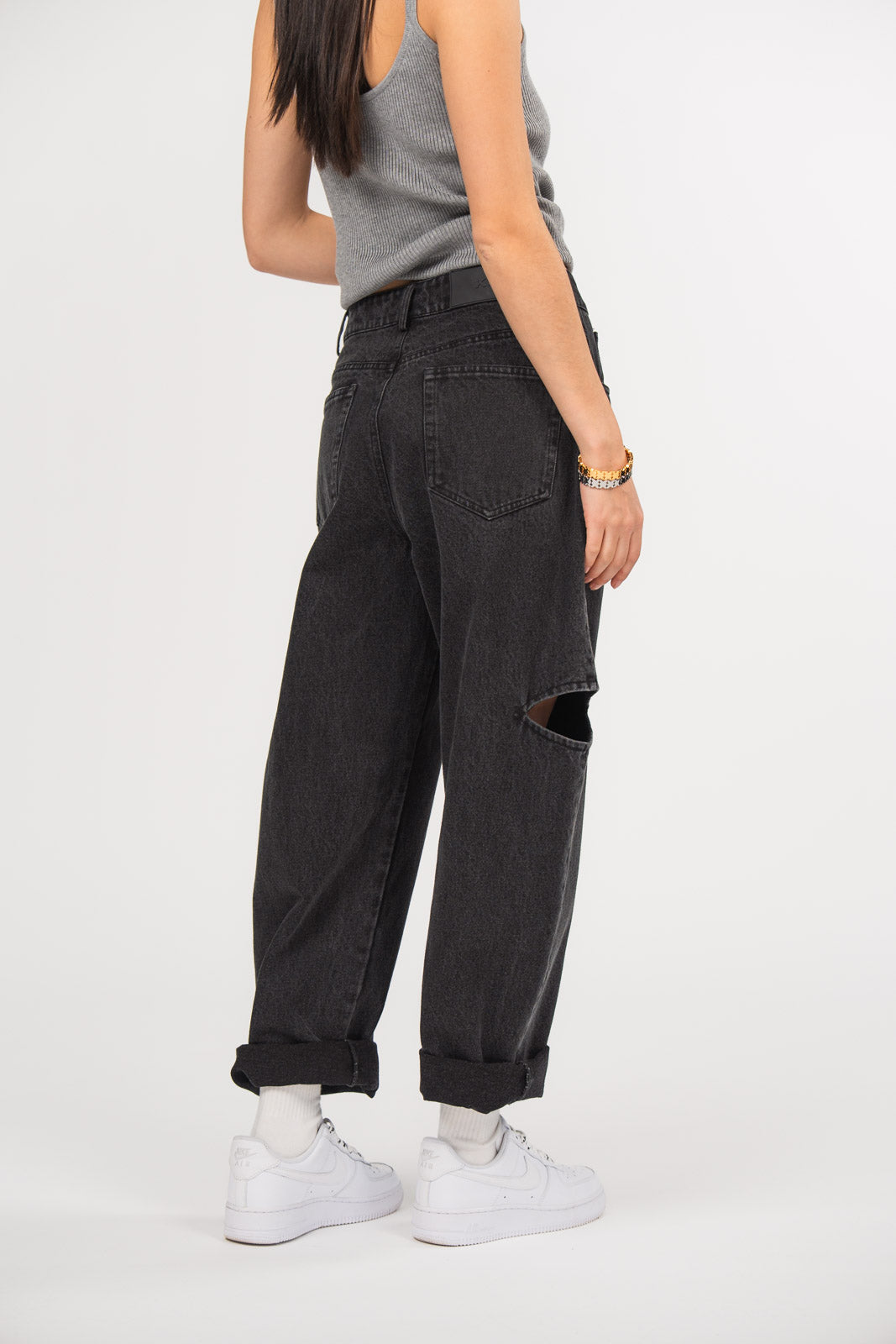Ein Model, von hinten gezeigt, trägt eine schwarze Cut-out-Jeans aus der No Hustle Culture Kollektion mit umgeschlagenem Saum, kombiniert mit einem grauen Tanktop, weißen Socken, weißen Sneakern und goldenem Armschmuck