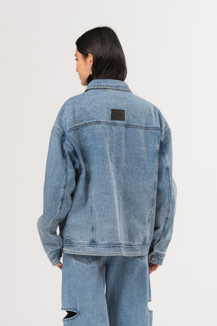 Ein Model, von hinten gezeigt, trägt eine blaue Denimjacke aus der No Hustle Culture Kollektion mit kleinem dunklem Logo-Patch im Nackenbereich, kombiniert mit einer blauen Cut-out-Jeans aus der No Hustle Culture Kollektion
