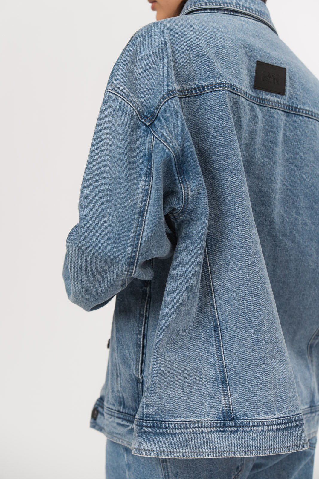 Ein Model, von hinten gezeigt, trägt eine blaue Denimjacke aus der No Hustle Culture Kollektion mit kleinem dunklem Logo-Patch im Nackenbereich
