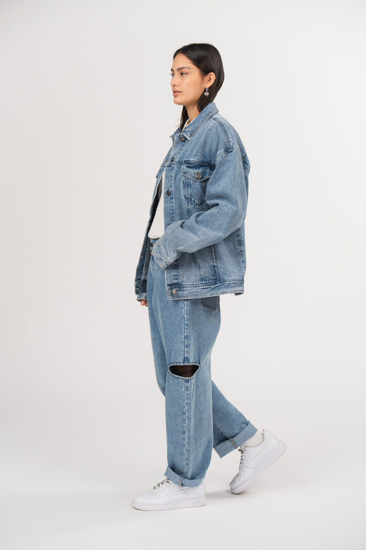 Ein Model in Seitenansicht trägt eine blaue Denimjacke und eine blaue Cut-out-Jeans aus der No Hustle Culture Kollektion, kombiniert mit einem weißen Strick-Polo und weißen Sneakern