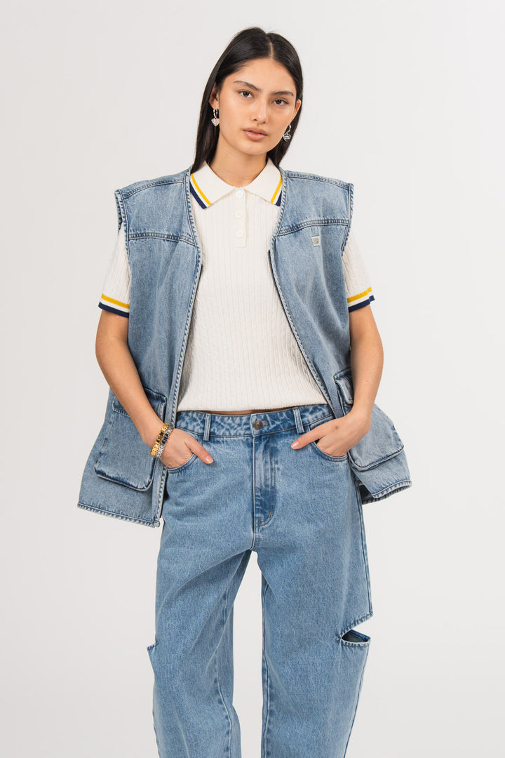 Ein Model trägt eine blaue Denimweste aus der No Hustle Culture Kollektion mit Reißverschluss, aufgesetzten Taschen und kleinem Logo-Patch auf der Brust, kombiniert mit einer blauen Cut-Out-Jeans aus der No Hustle Culture Kollektion und einem offwhite Strick-Poloshirt mit farblich abgesetztem Kragen.