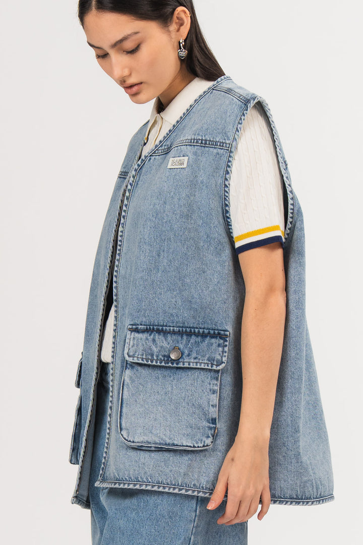 Ein Model in Seitenansicht trägt eine blaue Denimweste aus der No Hustle Culture Kollektion mit Reißverschluss, aufgesetzter Tasche und kleinem Logo-Patch auf der Brust, kombiniert mit einer blauen Jeans und einem offwhite Strick-Poloshirt