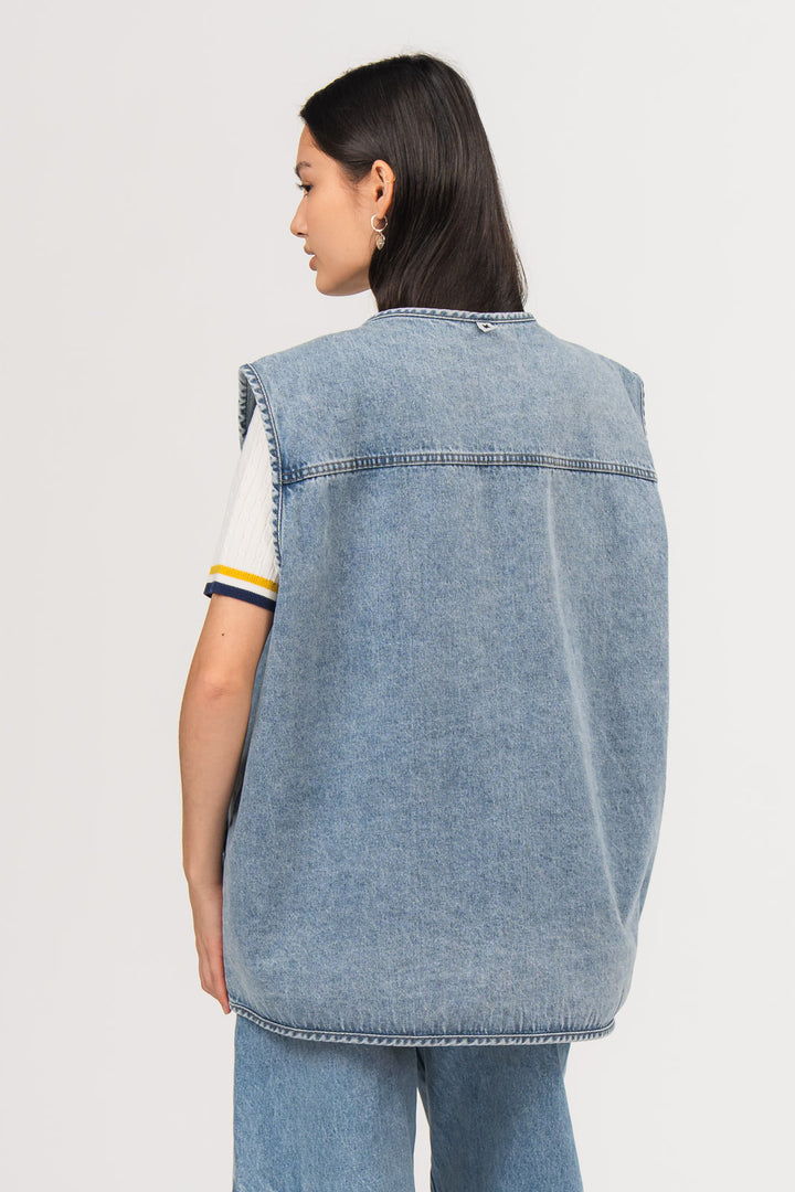 Ein Model, von hinten gezeigt, trägt eine blaue Denimweste aus der No Hustle Culture Kollektion mit kleinem Logo-Flag im Nackenbereich, kombiniert mit einer blauen Jeans aus der No Hustle Culture Kollektion und einem offwhite Strick-Poloshirt
