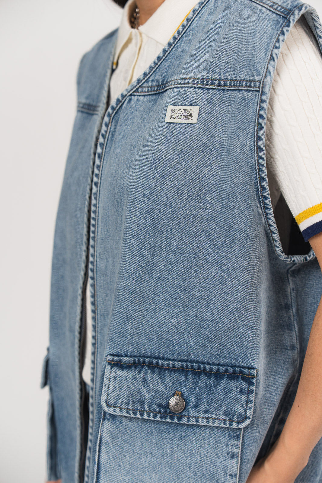Detailansicht einer blauen Denimweste aus der No Hustle Culture Kollektion mit kleinem Logo-Patch auf der Brust und aufgesetzter Tasche, kombiniert mit einem offwhite Strick-Poloshirt