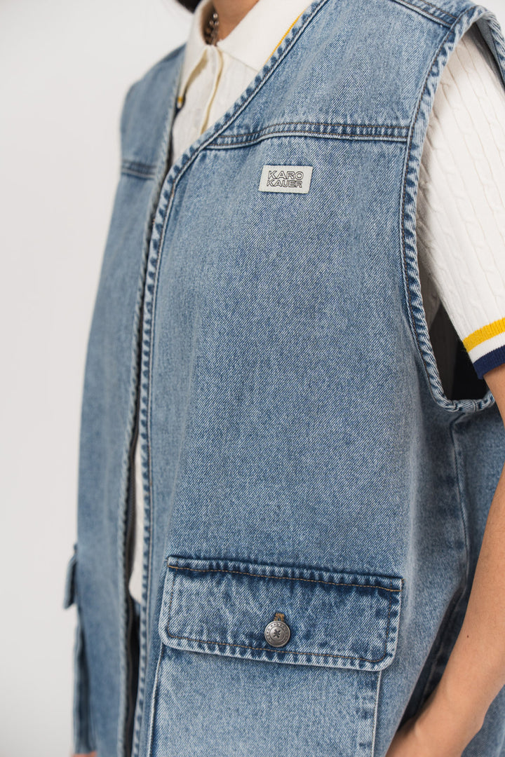 Detailansicht einer blauen Denimweste aus der No Hustle Culture Kollektion mit kleinem Logo-Patch auf der Brust und aufgesetzter Tasche, kombiniert mit einem offwhite Strick-Poloshirt