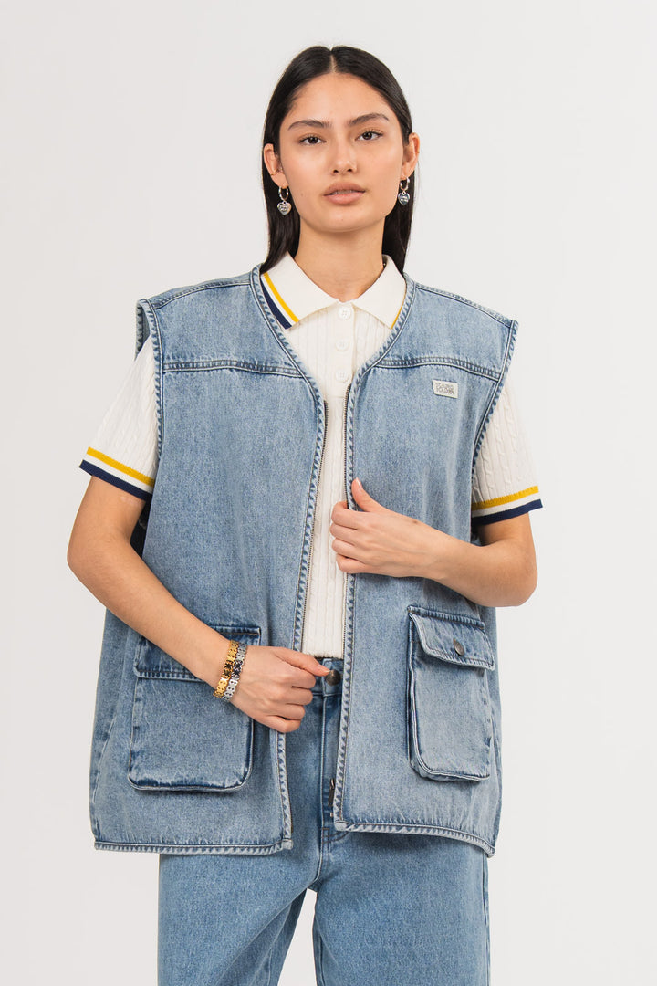Ein Model trägt eine blaue Denimweste aus der No Hustle Culture Kollektion mit Reißverschluss, aufgesetzten Taschen und kleinem Logo-Patch auf der Brust, kombiniert mit einer blauen Jeans aus der No Hustle Culture Kollektion und einem offwhite Strick-Poloshirt mit farblich abgesetztem Kragen