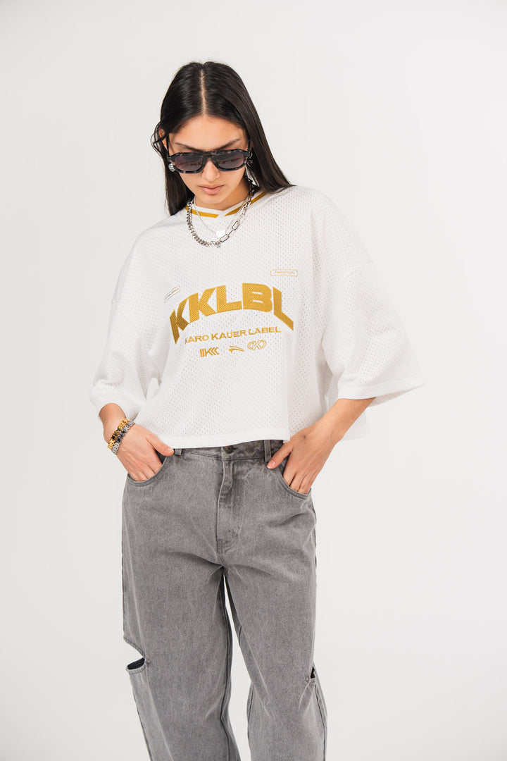 Ein Model trägt ein weißes cropped Jersey  mit großem gesticktem goldenem „KKLBL“-Logo auf der Brust, kombiniert mit einer grauen Cut-out-Jeans aus der No Hustle Culture Kollektion, schwarzer Sonnenbrille und silberfarbenem Schmuck