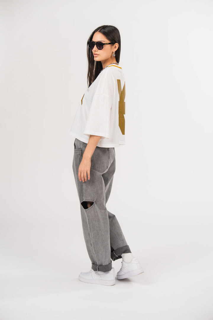 Ein Model in Seitenansicht trägt ein weißes cropped Jersey und eine graue Cut-out-Jeans aus der No Hustle Culture Kollektion, kombiniert mit weißen Sneakern und schwarzer Sonnenbrille