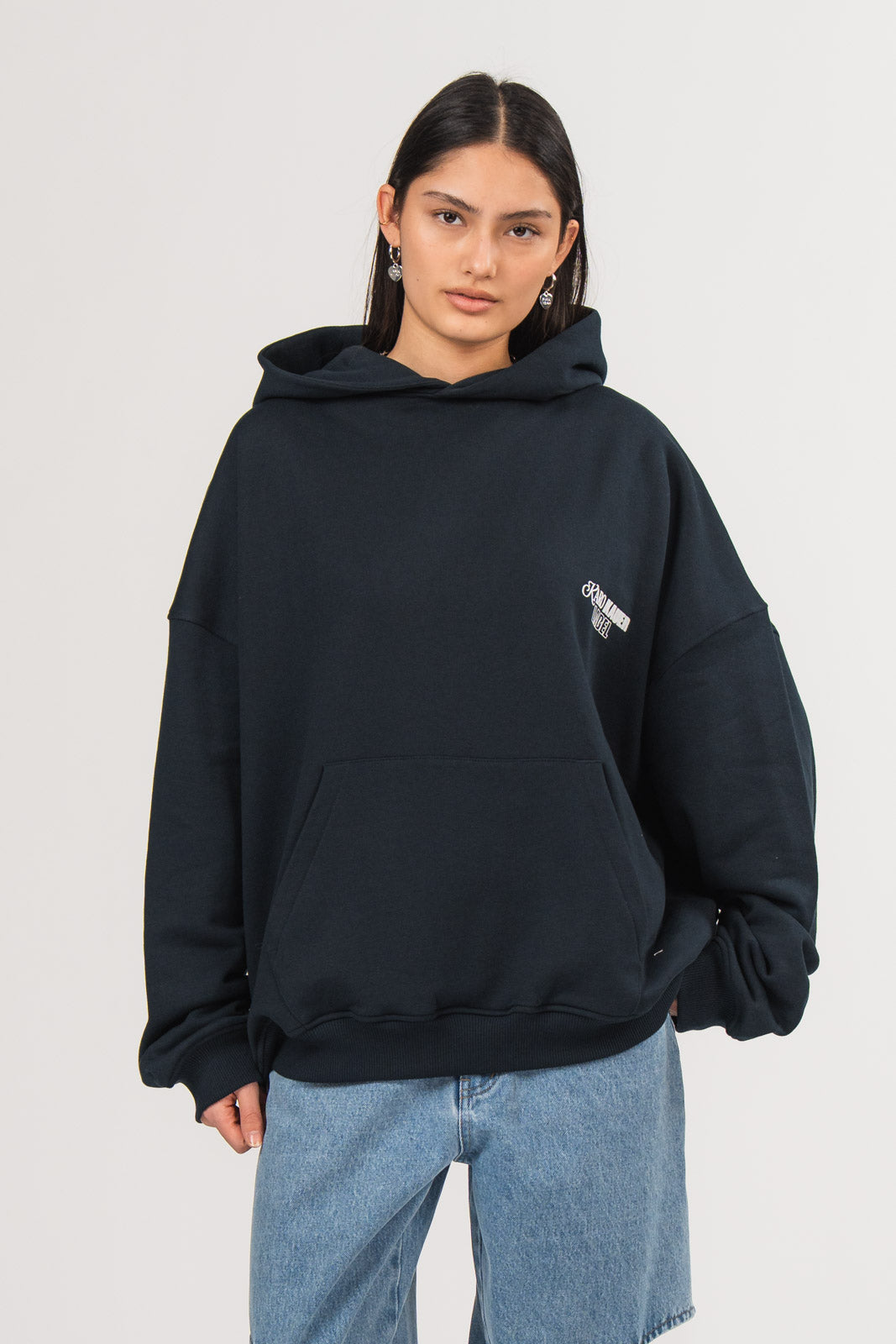 Ein Model trägt einen dunkelblauen Oversize-Hoodie mit Kapuze, aufgesetzter Tasche und kleinem weißen Brustprint, kombiniert mit einer hellblauen Cut-out-Jeans, aus der No Hustle Culture Kollektion