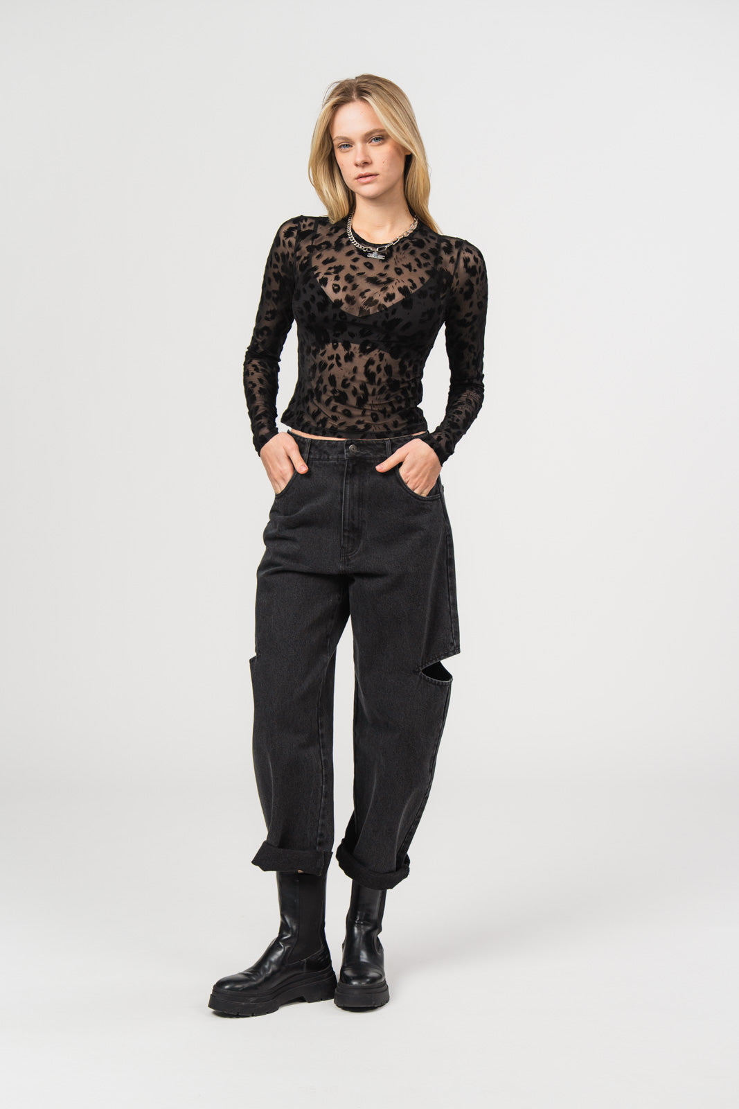 Eine Frau trägt ein transparentes schwarzes Mesh-Longsleeve mit samtigem Leopardenmuster, darunter ein schwarzes Bustier-Top, kombiniert mit einer schwarzen Cut-out-Hose aus der No Hustle Culture Kollektion, silberfarbener Kette und schwarzen Boots