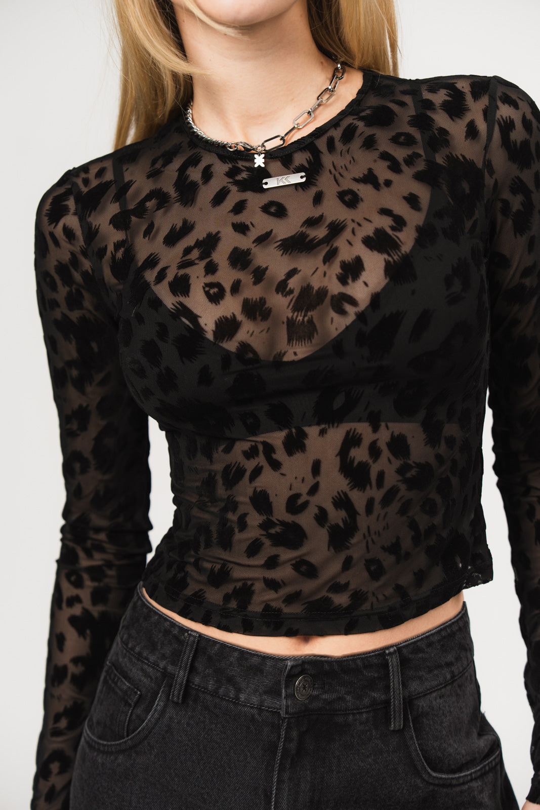 Oberkörperansicht einer Frau in einem transparenten schwarzen Mesh-Longsleeve mit samtigem Leopardenmuster, darunter ein schwarzes Bustier-Top, kombiniert mit dunkler Jeans und silberfarbener Kette