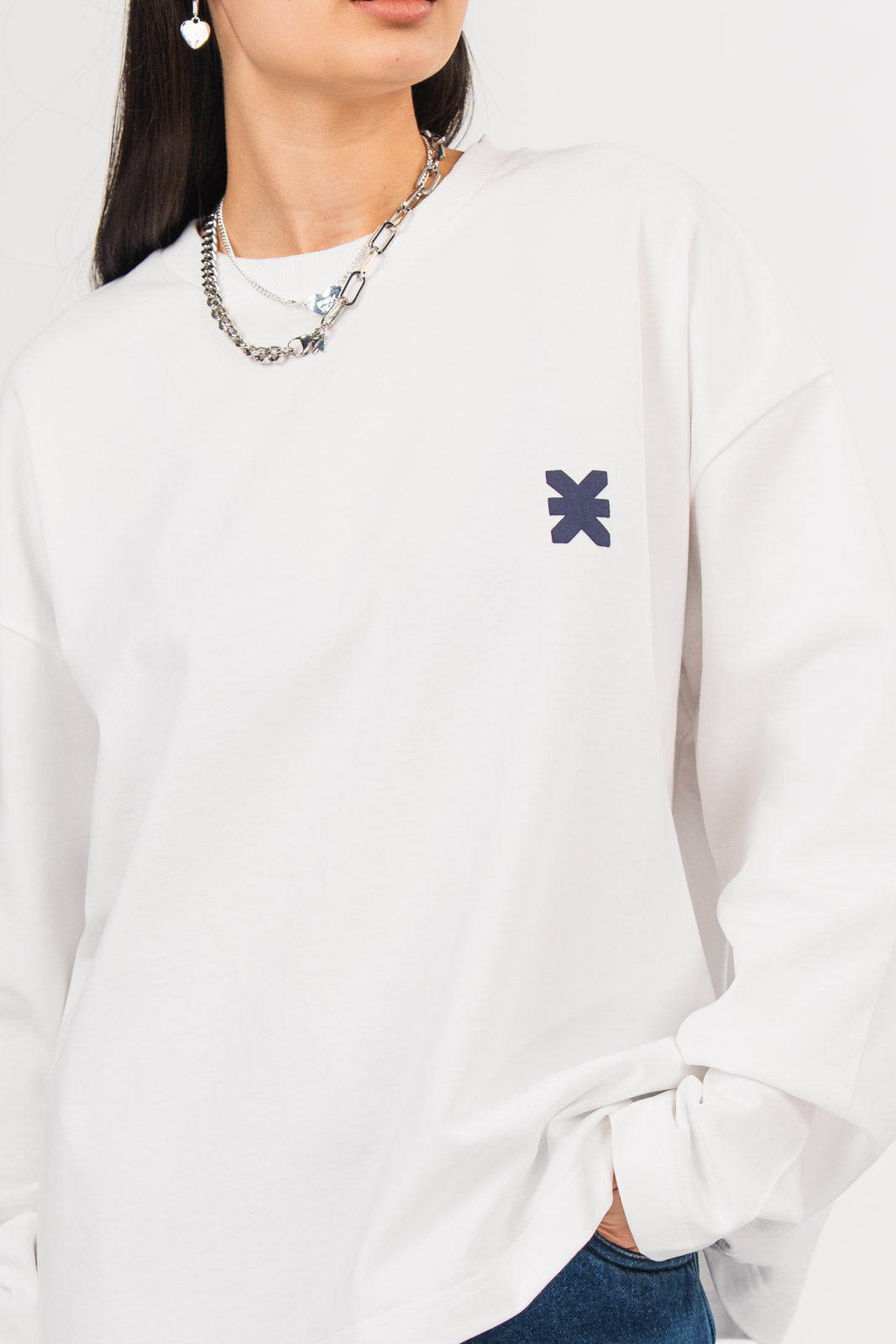 Detailansicht eines weißen Longsleeves mit Rundhalsausschnitt und kleinem dunkelblauen Logo-Print auf der Brust, kombiniert mit silberfarbenem Schmuck