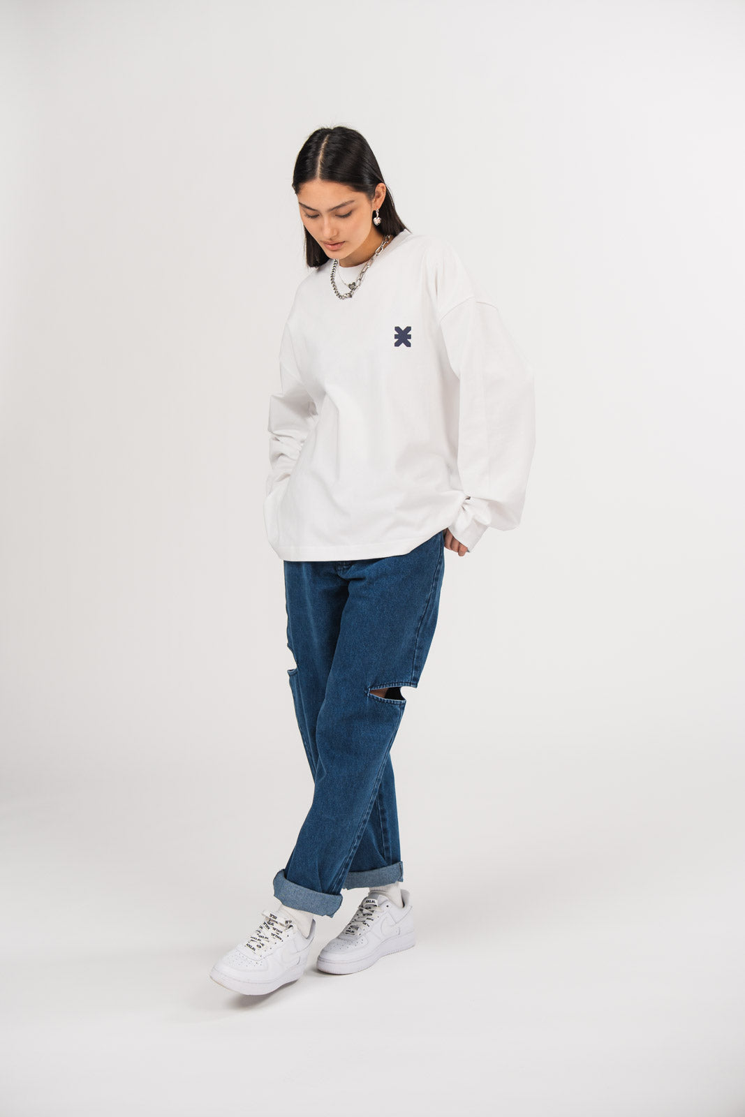 Ein Model in Ganzkörperansicht trägt ein weißes Longsleeve mit Rundhalsausschnitt und kleinem dunkelblauen Logo-Print auf der Brust, kombiniert mit einer dunkelblauen Cut-out-Jeans aus der No Hustle Culture Kollektion, weißen Sneakern und silberfarbenem Schmuck.