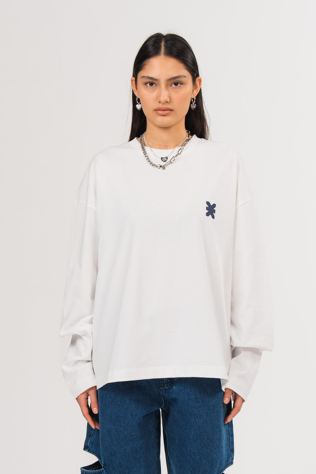 Ein Model trägt ein weißes Longsleeve mit Rundhalsausschnitt und kleinem dunkelblauen Logo-Print auf der Brust, kombiniert mit einer dunkel blauen Cut-Out-Jeans aus der No Hustle Culture Kollektion und silberfarbenem Schmuck