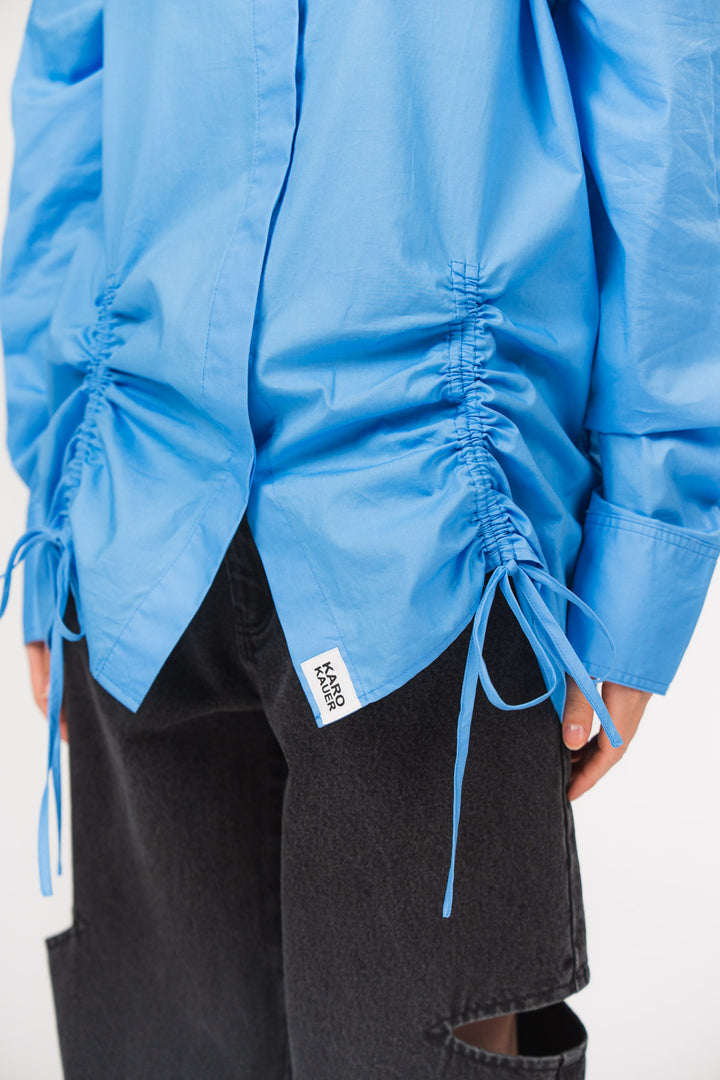 Detailansicht einer hellblauen Bluse mit seitlichen Raffungen, Bindebändern und kleinem Weblabel am Saum, kombiniert mit einer schwarzen Cut-out-Jeans aus der No Hustle Culture Kollektion