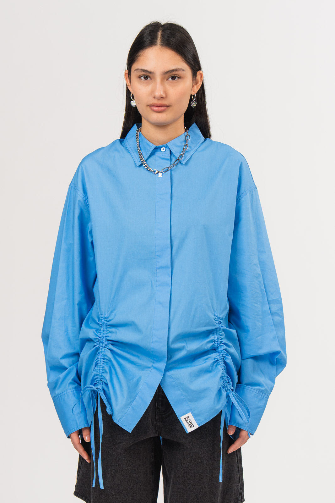 Ein Model trägt eine hellblaue Bluse mit Kragen, verdeckter Knopfleiste, seitlichen Raffungen, Bindebändern und kleinem Weblabel am Saum, kombiniert mit einer schwarzen Cut-out-Jeans aus der No Hustle Culture Kollektion und silberfarbenem Schmuck
