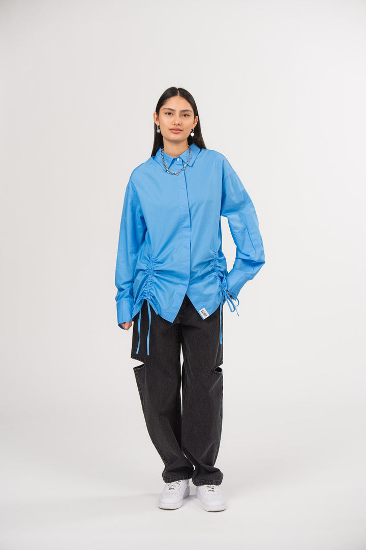 Ein Model trägt eine hellblaue Bluse mit Kragen, verdeckter Knopfleiste, seitlichen Raffungen, Bindebändern und kleinem Weblabel am Saum, kombiniert mit einer schwarzen Cut-out-Jeans aus der No Hustle Culture Kollektion, silberfarbenem Schmuck und weißen Sneakern