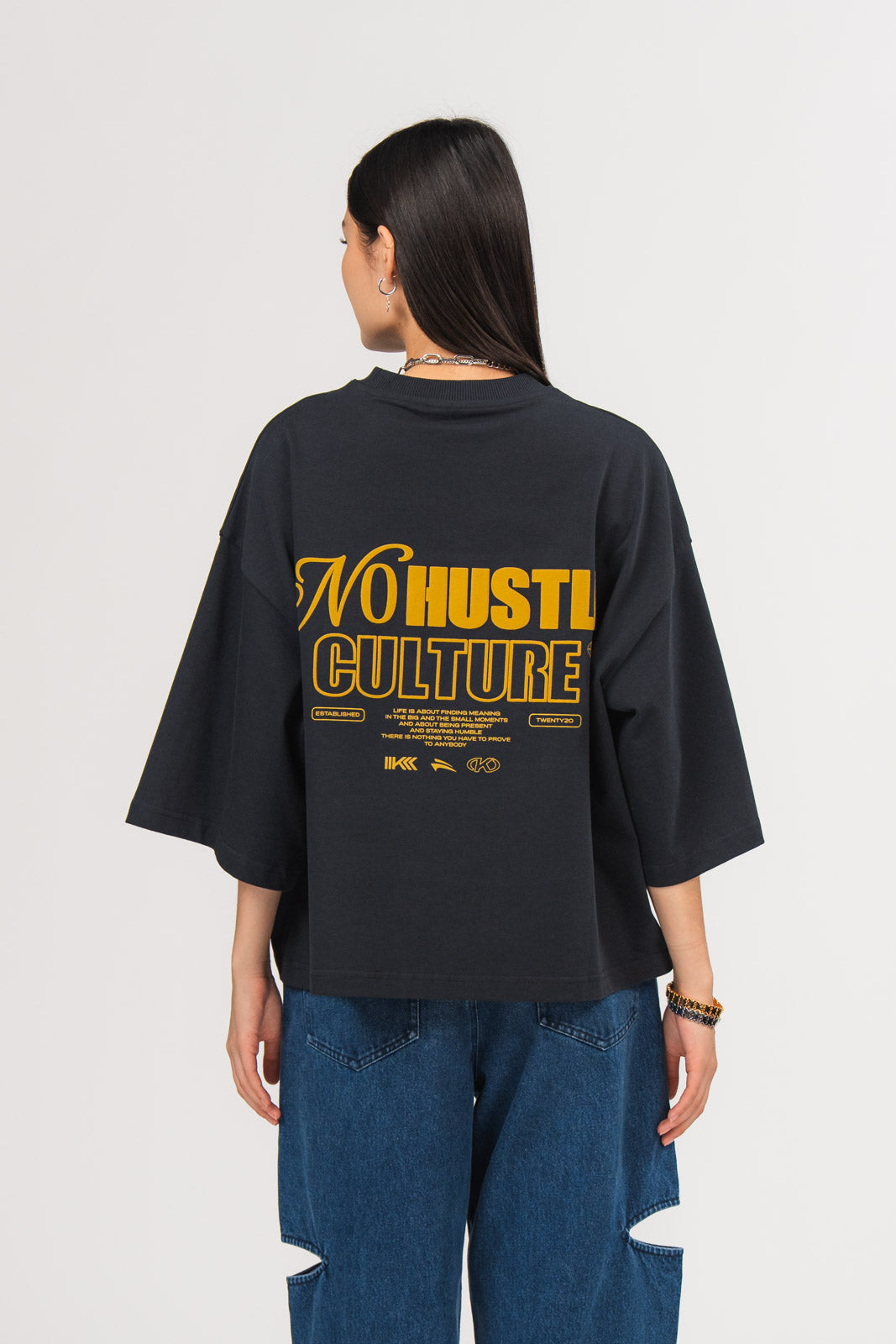 Ein Model, von hinten gezeigt, trägt ein dunkelblaues oversized T-Shirt mit großem gelbem Rückenprint aus dem Schriftzug „No Hustle Culture“, kleinem Textblock und Logo-Elementen, kombiniert mit einer dunkelblauen Cut-out-Jeans aus der No Hustle Culture Kollektion