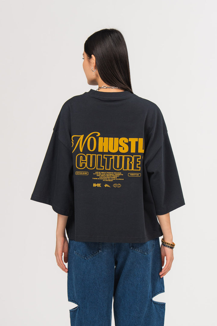 Ein Model, von hinten gezeigt, trägt ein dunkelblaues oversized T-Shirt mit großem gelbem Rückenprint aus dem Schriftzug „No Hustle Culture“, kleinem Textblock und Logo-Elementen, kombiniert mit einer dunkelblauen Cut-out-Jeans aus der No Hustle Culture Kollektion