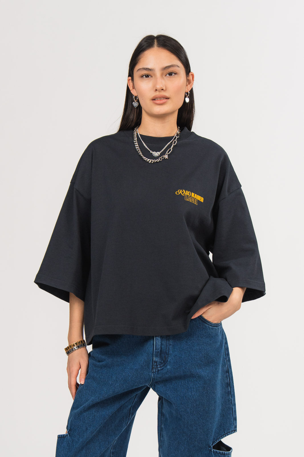Ein Model trägt ein dunkelblaues oversized T-Shirt aus der No Hustle Culture Kollektion mit kleinem gelbem „KARO KAUER LABEL“-Print auf der Brust, kombiniert mit einer dunkelblauen Cut-out-Jeans aus der No Hustle Culture Kollektion und silberfarbenem Schmuck