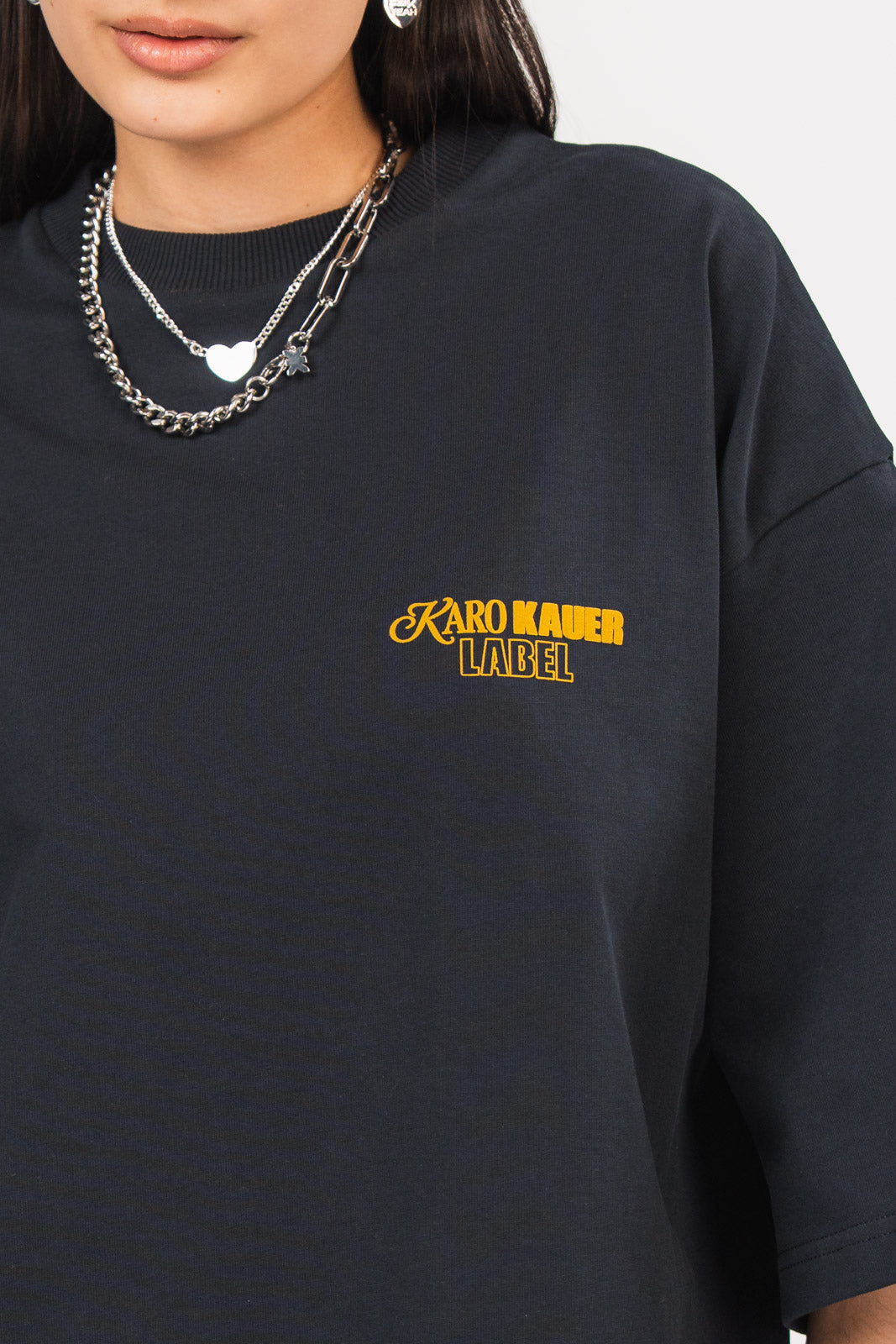 Detailansicht eines dunkelblauen oversized T-Shirts aus der No Hustle Culture Kollektion mit kleinem gelbem „KARO KAUER LABEL“-Print auf der Brust, kombiniert mit silberfarbenem Schmuck.