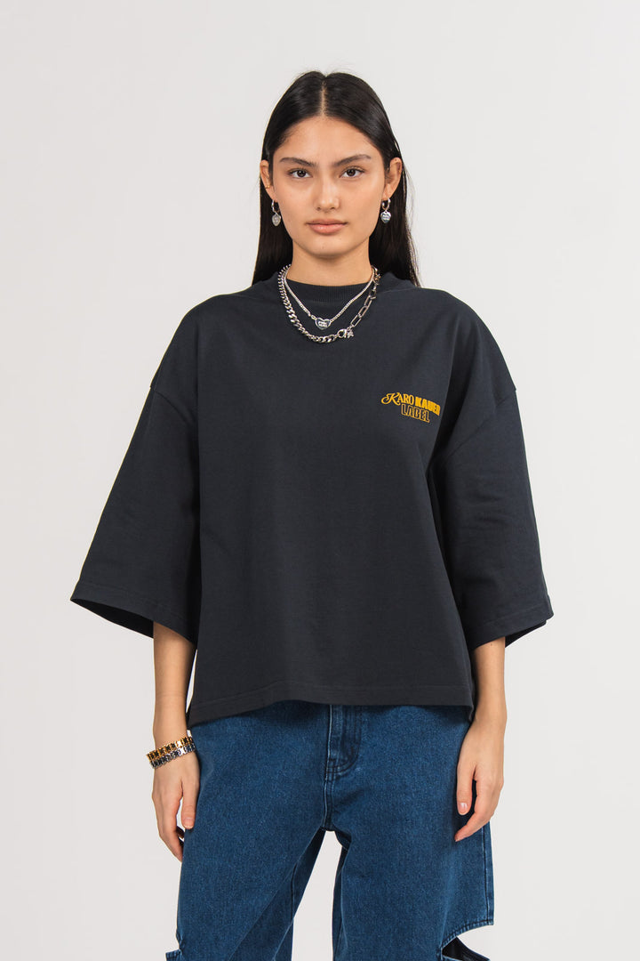 Ein Model trägt ein dunkelblaues oversized T-Shirt mit kleinem gelbem „KARO KAUER LABEL“-Print auf der Brust, kombiniert mit einer dunkelblauen Cut-out-Jeans aus der No Hustle Culture Kollektion und silberfarbenem Schmuck