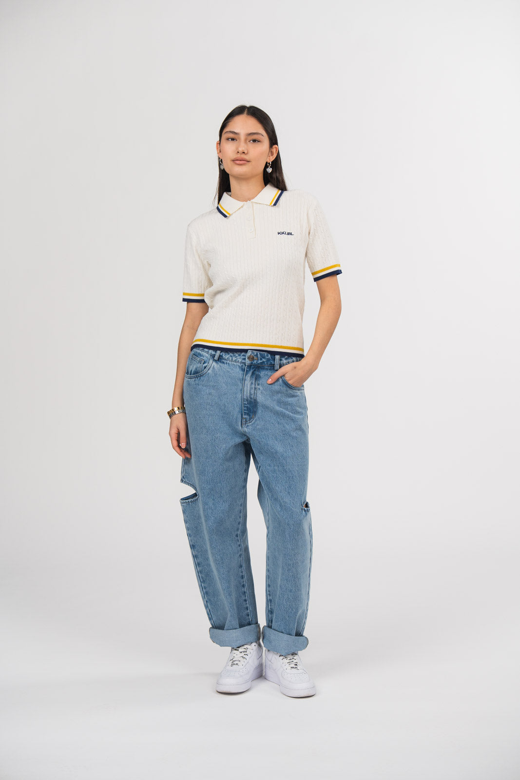 Ein Model in Ganzkörperansicht trägt ein offwhite Strick-Poloshirt mit weißem Kragen, gelben und dunkelblauen Kontraststreifen sowie kleinem dunkelblauen, gestickten „KKLBL“-Logo auf der Brust, kombiniert mit einer blauen Cut-out-Jeans aus der No Hustle Culture Kollektion und weißen Sneakern