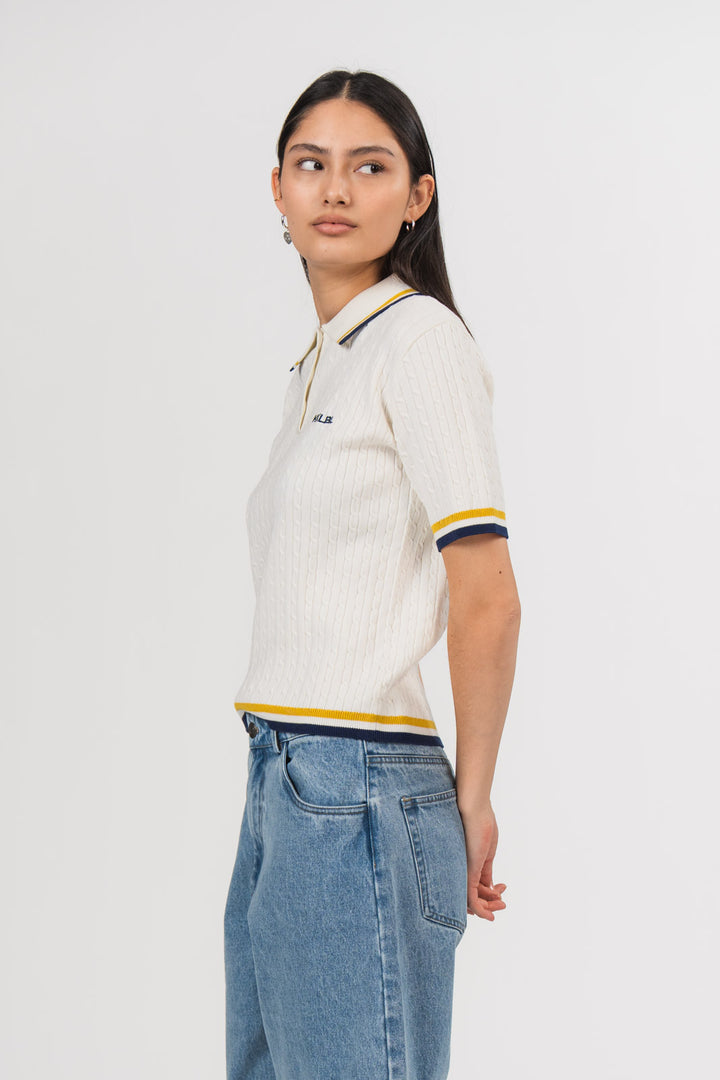 Ein Model in Seitenansicht trägt ein offwhite Strick-Poloshirt mit weißem Kragen, gelben und dunkelblauen Kontraststreifen sowie kleinem dunkelblauen, gestickten „KKLBL“-Logo auf der Brust, kombiniert mit einer hellblauen Jeans aus der No Hustle Culture Kollektion