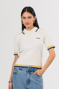 Ein Model in Ganzkörperansicht trägt ein offwhite Strick-Poloshirt mit weißem Kragen, gelben und dunkelblauen Kontraststreifen sowie kleinem dunkelblauen, gestickten „KKLBL“-Logo auf der Brust, kombiniert mit einer blauen Cut-out-Jeans aus der No Hustle Culture Kollektion 
