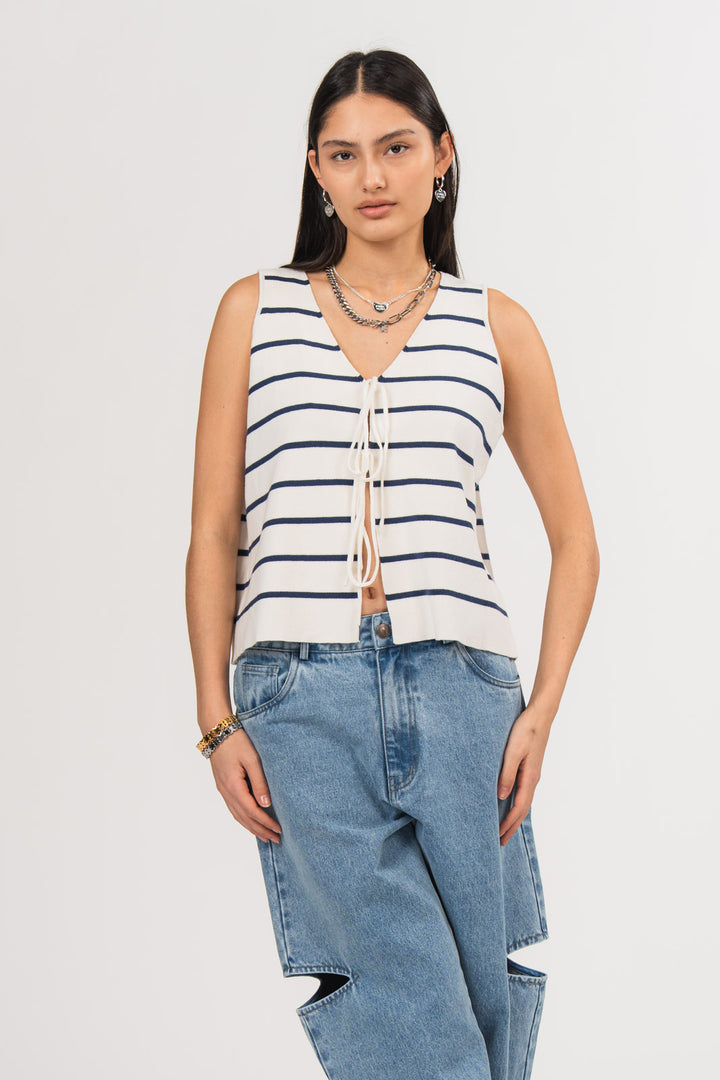 Ein Model in Ganzkörperansicht trägt eine gestrickte Weste mit dunkelblauen Streifen und mehreren Schleifen-Details auf der Vorderseite, kombiniert mit einer blauen Cut-out-Jeans aus der No Hustle Culture Kollektion