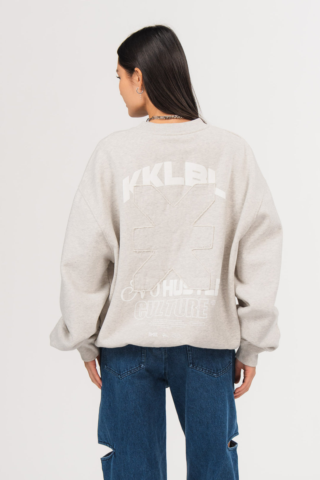 Ein Model, von hinten gezeigt, trägt ein hellgraues Oversize-Sweatshirt aus der No Hustle Culture Kollektion mit großem Rückenprint aus weißem „KKLBL“-Schriftzug, weißem „No Hustle Culture“-Schriftzug und aufgesetztem farbgleichem Logo-Patch in der Mitte, kombiniert mit einer dunkel blauen Cutt-Out-Jeans aus der No Hustle Culture Kollektion.