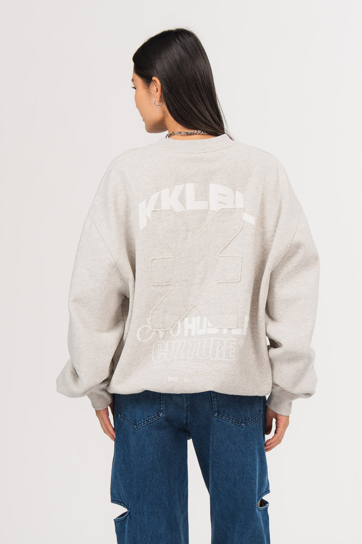 Ein Model, von hinten gezeigt, trägt ein hellgraues Oversize-Sweatshirt aus der No Hustle Culture Kollektion mit großem Rückenprint aus weißem „KKLBL“-Schriftzug, weißem „No Hustle Culture“-Schriftzug und aufgesetztem farbgleichem Logo-Patch in der Mitte, kombiniert mit einer dunkel blauen Cutt-Out-Jeans aus der No Hustle Culture Kollektion.