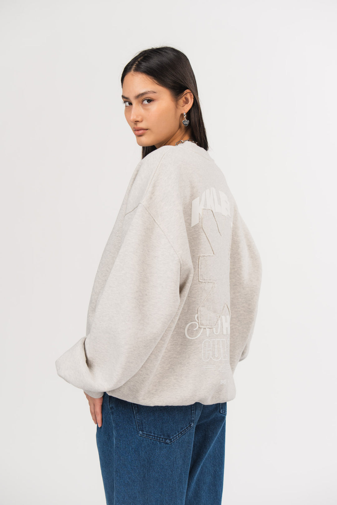 Ein Model in Seitenansicht trägt ein hellgraues Oversize-Sweatshirt mit großem weißem und farbgleichem Rückenprint sowie eine blaue Jeans aus der No Hustle Culture Kollektion, kombiniert mit silberfarbenen Ohrringen