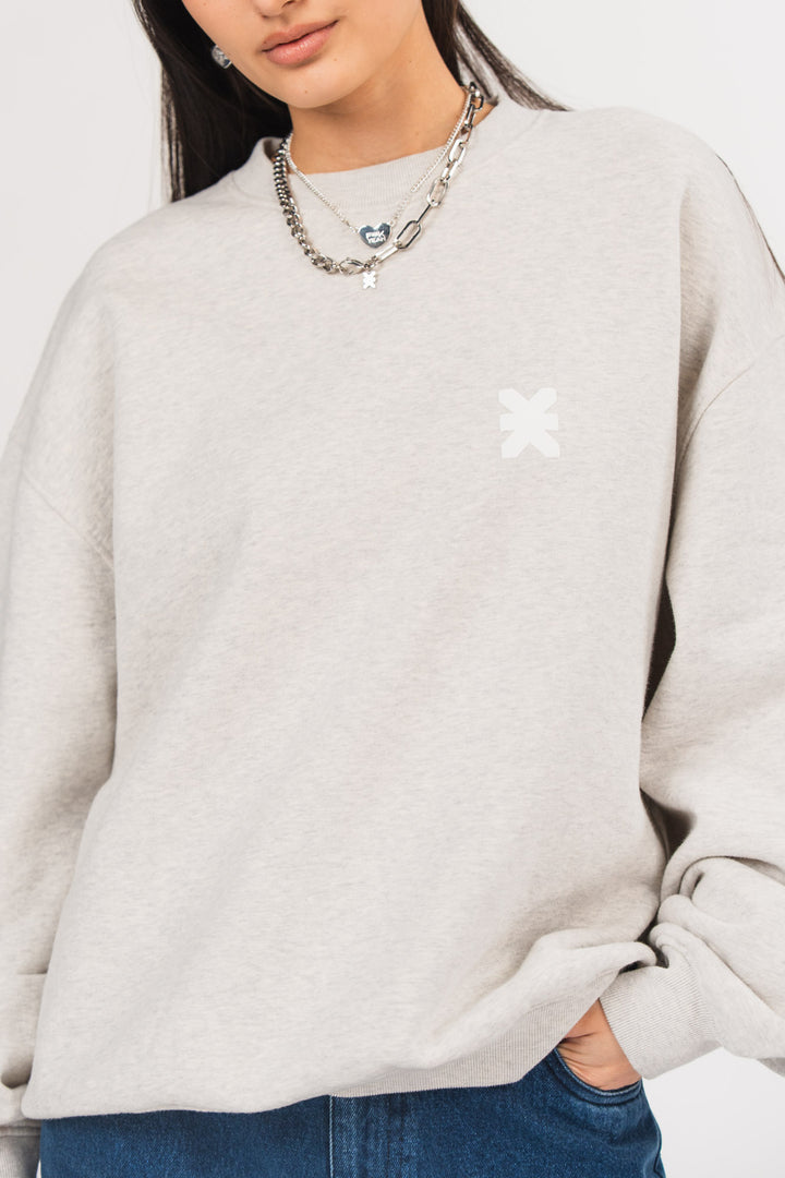 Ein Model trägt ein hellgraues Oversize-Sweatshirt mit Rundhalsausschnitt und kleinem tonalem Logo-Print auf der Brust, kombiniert mit einer blauen Jeans und silberfarbenem Schmuck