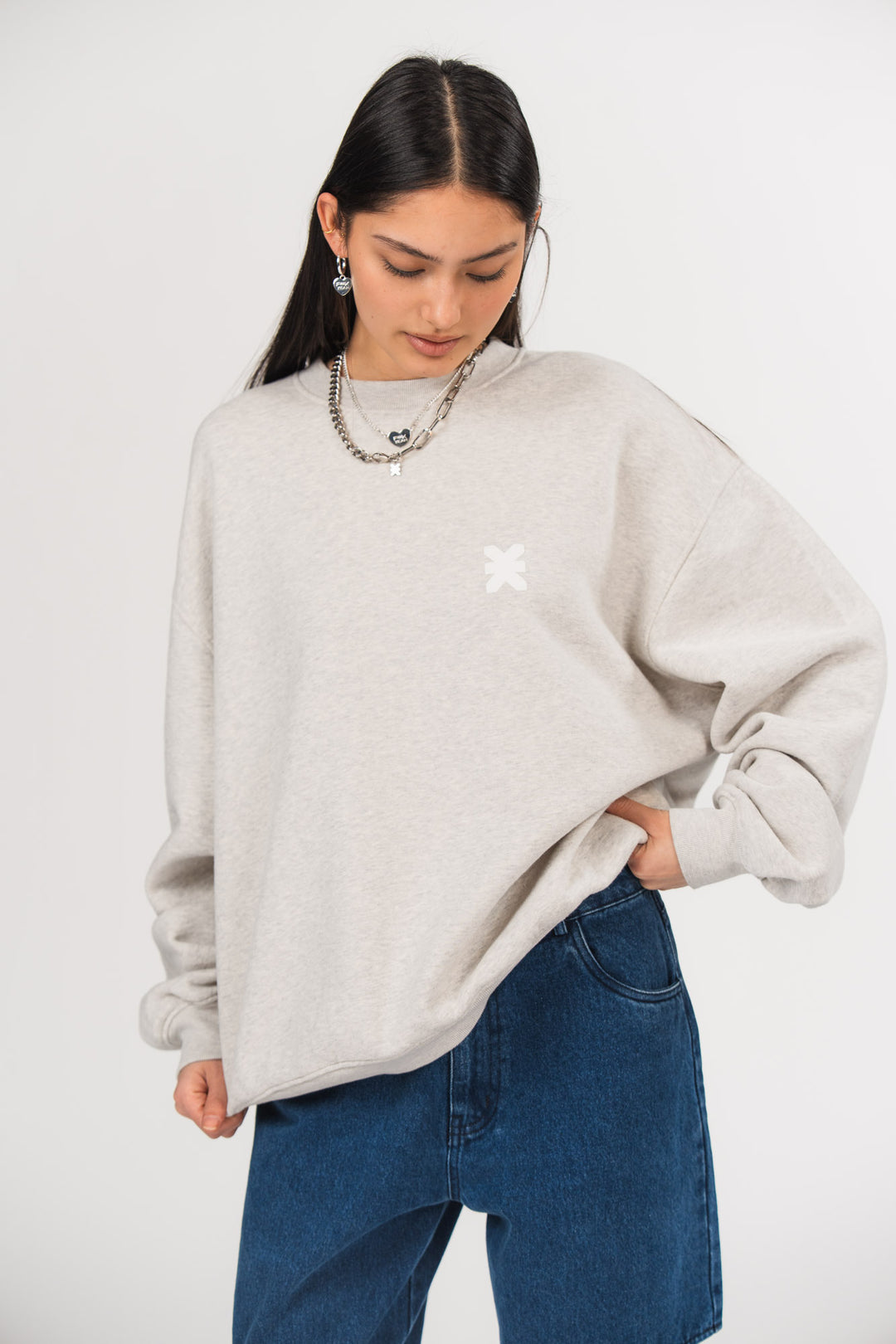 Ein Model trägt ein hellgraues Oversize-Sweatshirt mit Rundhalsausschnitt und kleinem weißen Logo-Print auf der Brust, kombiniert mit einer dunkel blauen Jeans, aus der No Hustle Culture Kollektion und silberfarbenem Schmuck