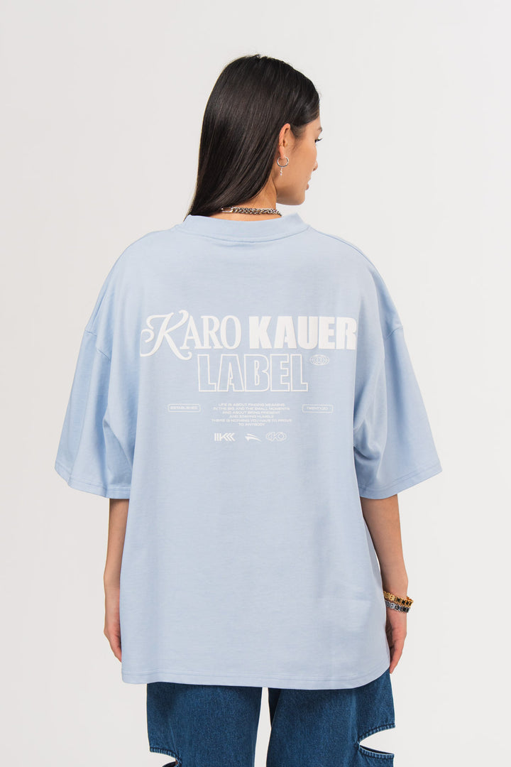 Ein Model, von hinten gezeigt, trägt ein hellblaues oversized T-Shirt aus der No Hustle Culture Kollektion mit großem weißem Rückenprint aus dem Schriftzug „KARO KAUER LABEL“, kleinem Textblock und Logo-Elementen, kombiniert mit einer dunkelblauen Cut-out-Jeans aus der No Hustle Culture Kollektion