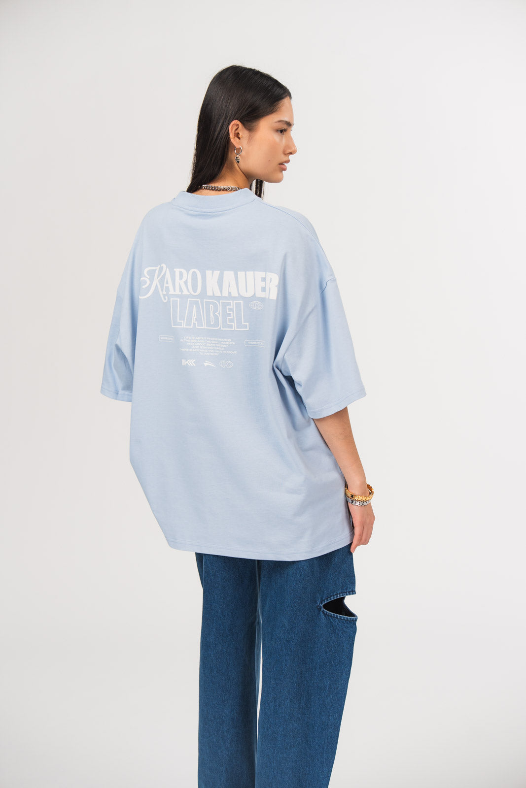 Ein Model in Seitenansicht trägt ein hellblaues oversized T-Shirt mit großem weißem Rückenprint aus dem Schriftzug „KARO KAUER LABEL“, kleinem Textblock und Logo-Elementen, kombiniert mit einer dunkelblauen Cut-out-Jeans aus der No Hustle Culture Kollektion