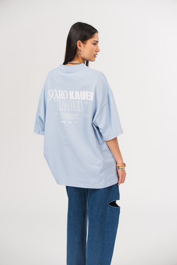 Ein Model in Seitenansicht trägt ein hellblaues oversized T-Shirt mit großem weißem Rückenprint aus dem Schriftzug „KARO KAUER LABEL“, kleinem Textblock und Logo-Elementen, kombiniert mit einer dunkelblauen Cut-out-Jeans aus der No Hustle Culture Kollektion