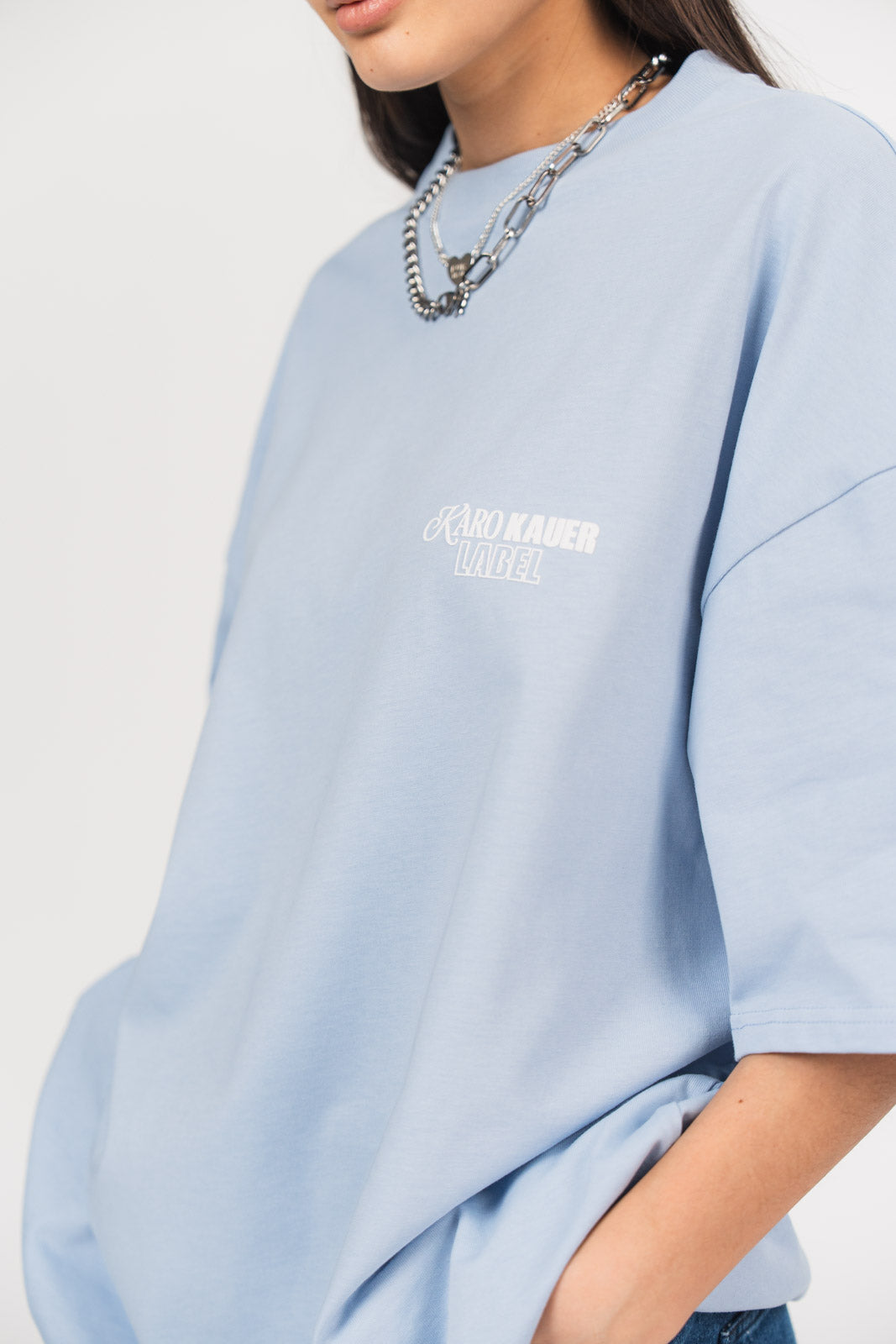 Detailansicht eines hellblauen oversized T-Shirts aus der No Hustle Culture Kollektion mit kleinem weißem „KARO KAUER LABEL“-Print auf der Brust, kombiniert mit silberfarbenem Schmuck