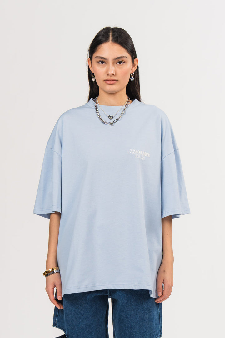 Ein Model trägt ein hellblaues oversized T-Shirt mit kleinem weißem „KARO KAUER LABEL“-Print auf der Brust, kombiniert mit einer dunkelblauen Cut-out-Jeans aus der No Hustle Culture Kollektion und silberfarbenem Schmuck