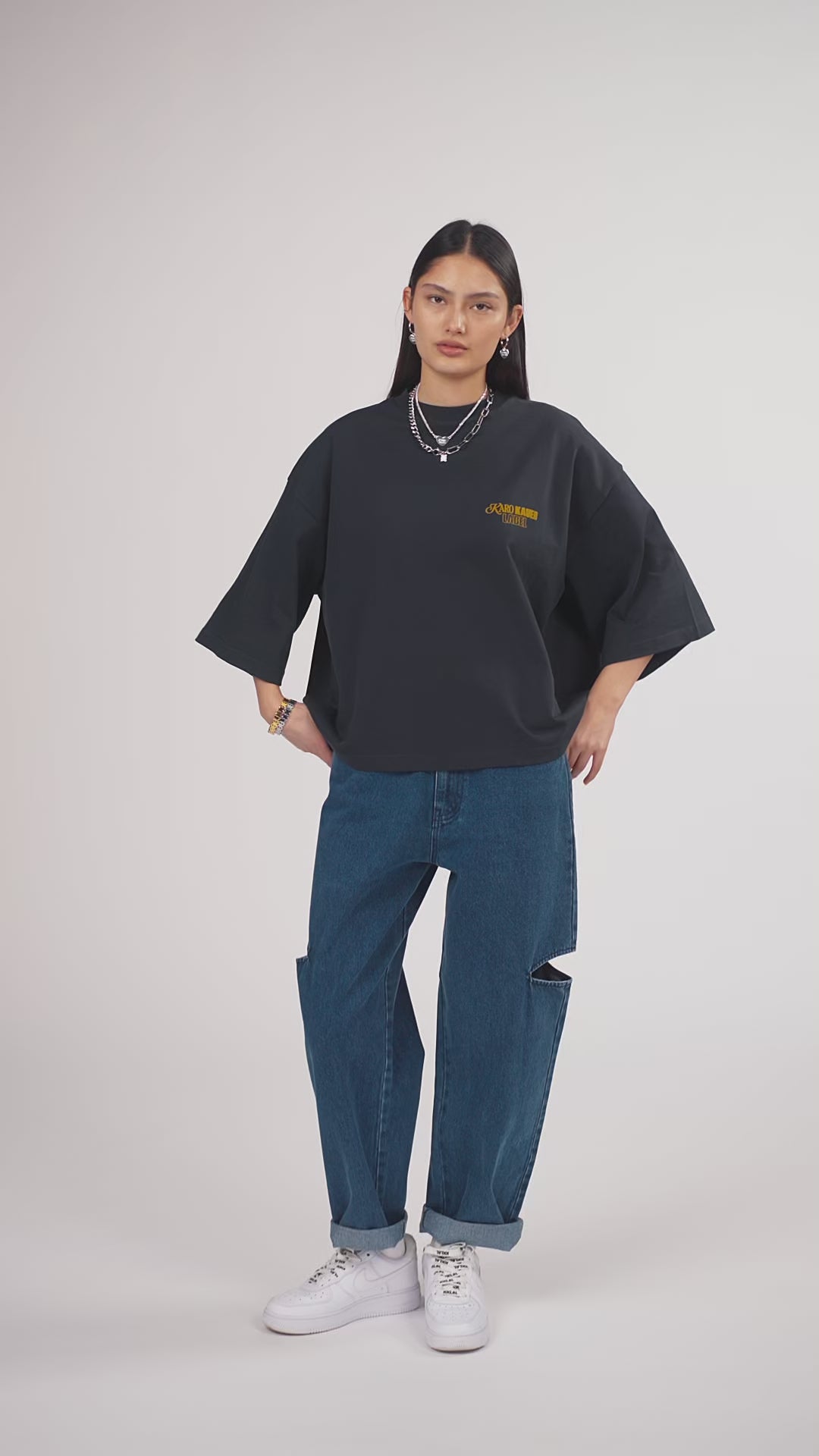 Ein Model in Ganzkörperansicht, das den Look in Bewegung aus verschiedenen Perspektiven zeigt, trägt ein hellgraues Oversize-Sweatshirt mit verschiedenen gelbfarbigen Prints, kombiniert mit einer blauen Cut-out-Jeans aus der No Hustle Culture Kollektion, weißen Sneakern und silberfarbenem Schmuck