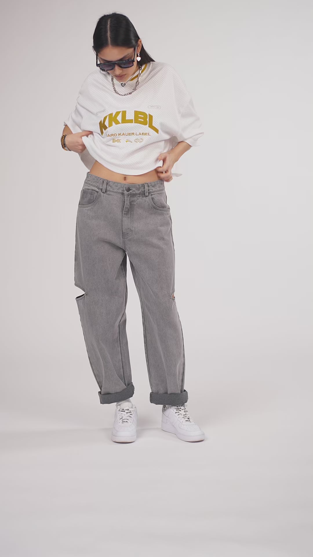 Ein Model in Ganzkörperansicht, das den Look in Bewegung aus verschiedenen Perspektiven zeigt, trägt ein weißes oversized Jersey-Shirt mit goldenem „KKLBL“-Print, kombiniert mit einer grauen Cut-out-Jeans aus der No Hustle Culture Kollektion, weißen Sneakern, Sonnenbrille und silberfarbenem Schmuck